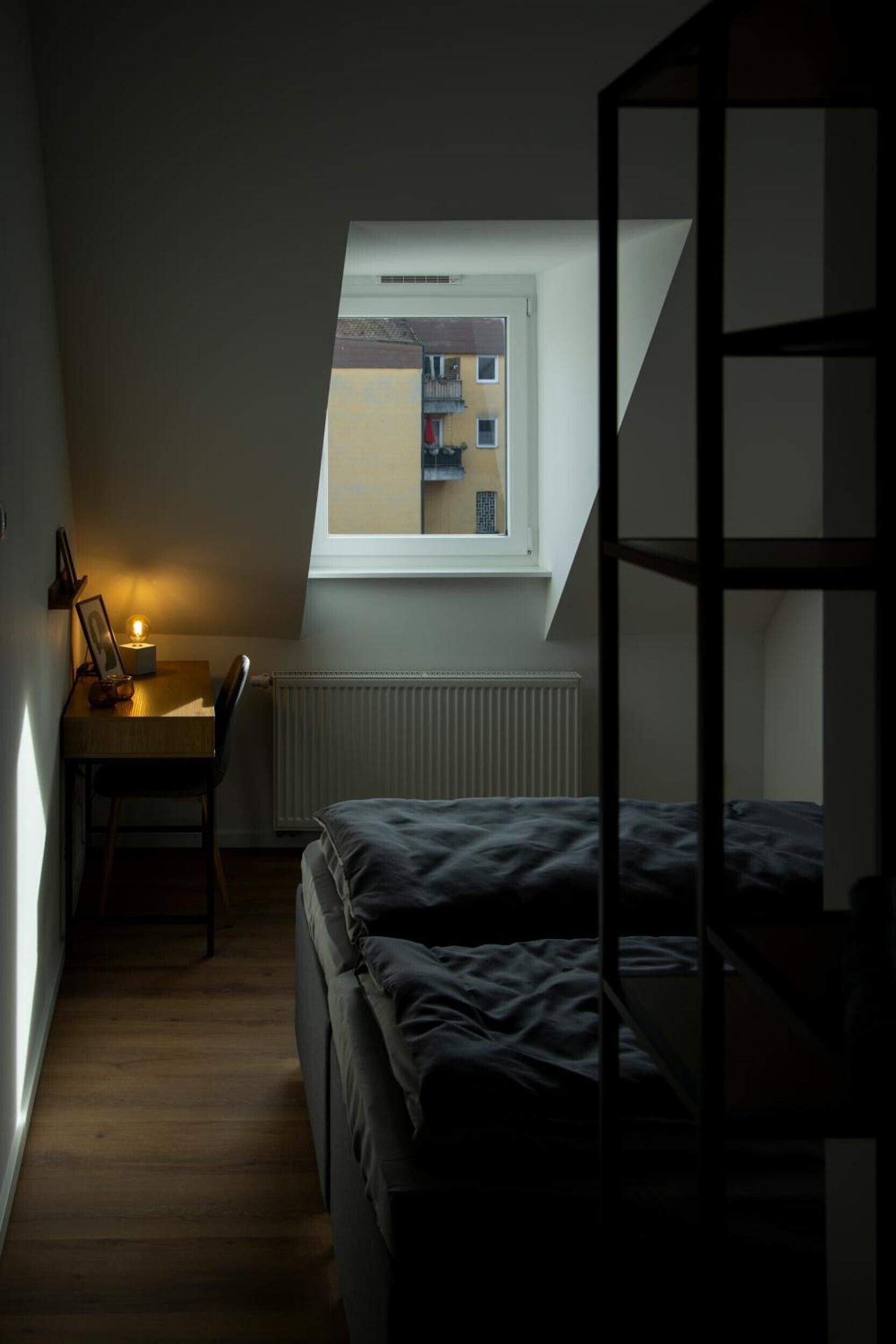 Ein kleiner, schwach beleuchteter Raum mit schräger Decke, einem Einzelbett, einem Schreibtisch mit Stuhl und einem Fenster mit Blick auf ein Gebäude.