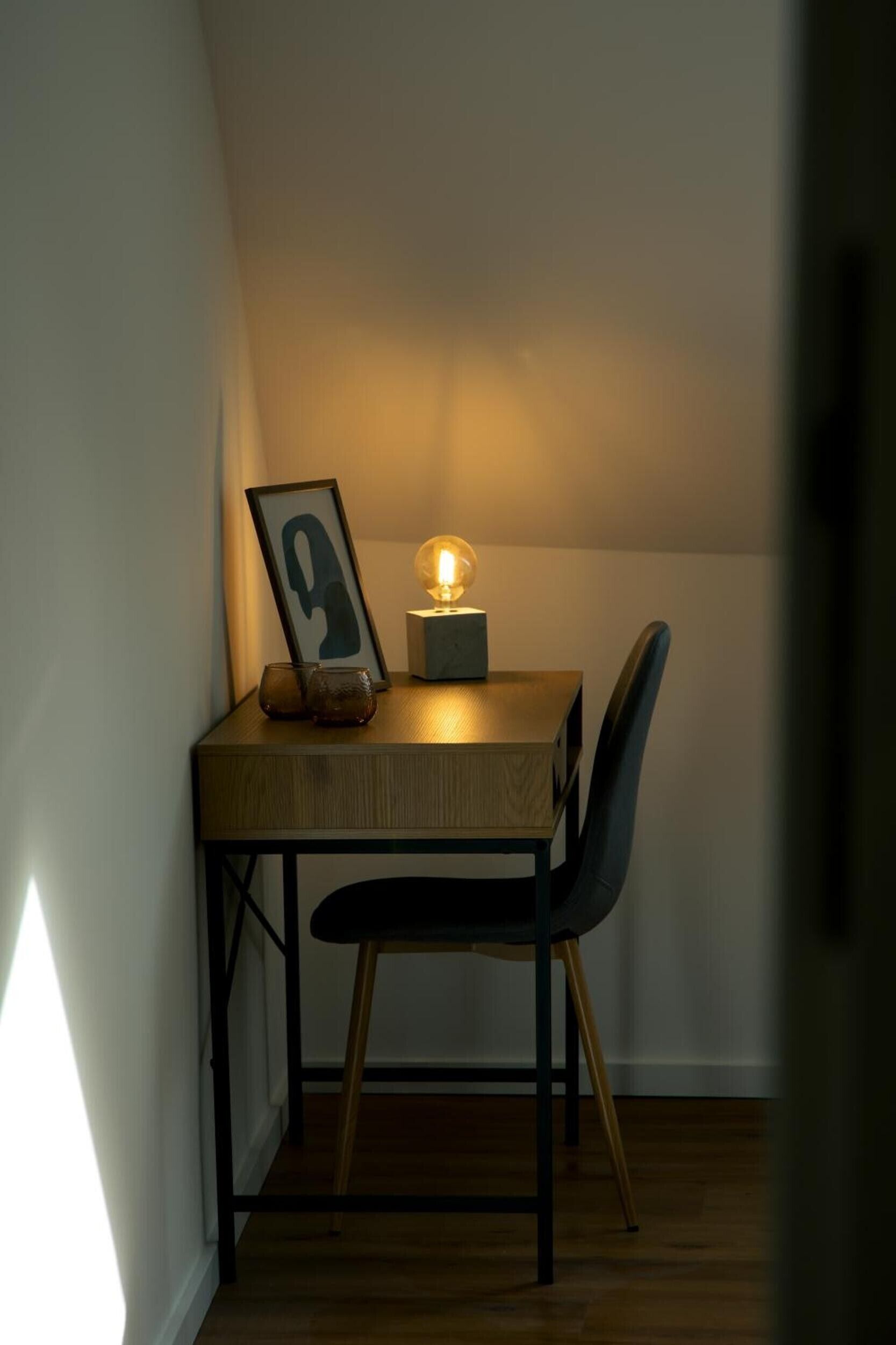 A corner room with a wooden table, a chair, a framed picture, and a table lamp.