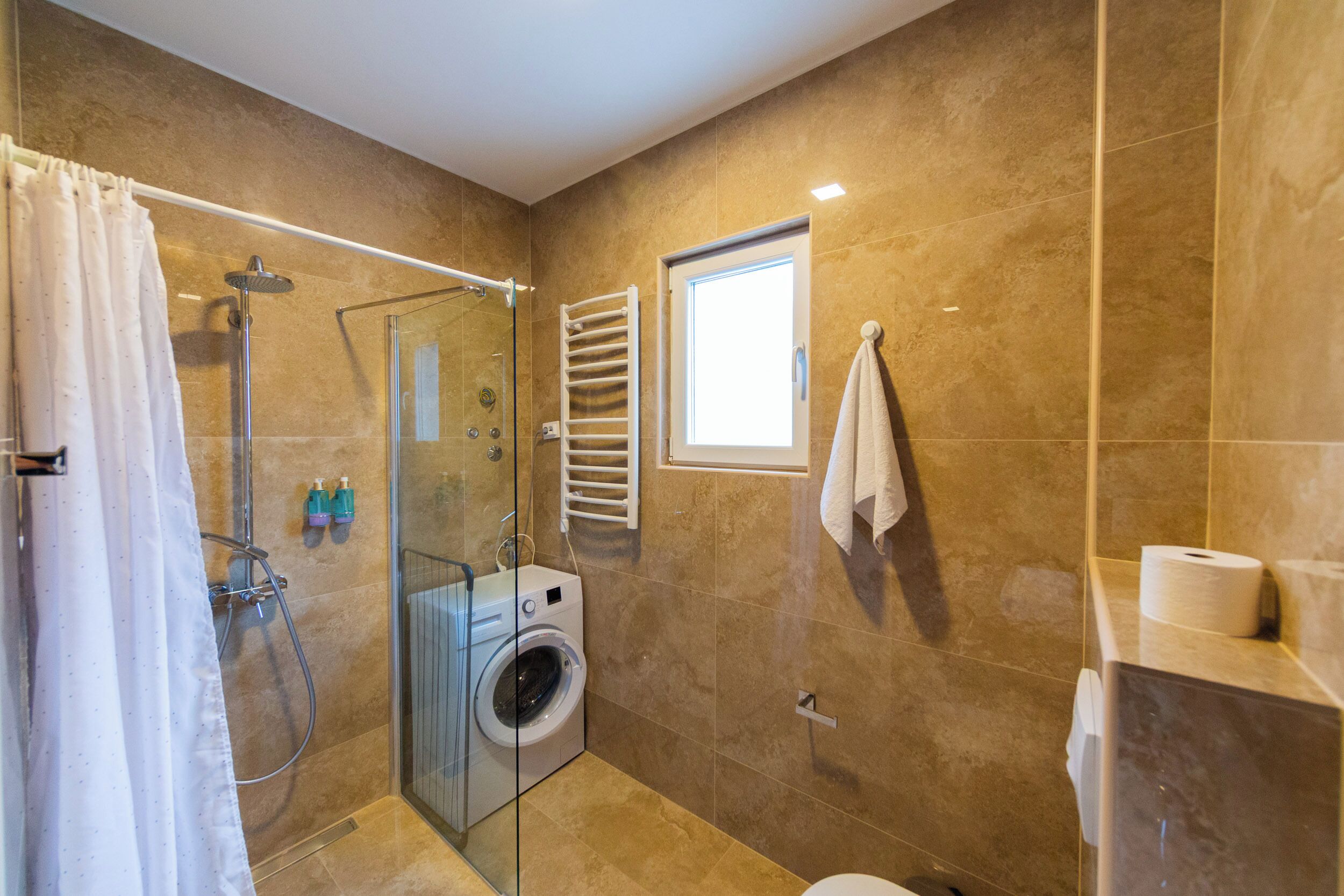 A modern bathroom with a glass-enclosed shower, a built-in washing machine, a toilet, and a towel rack.