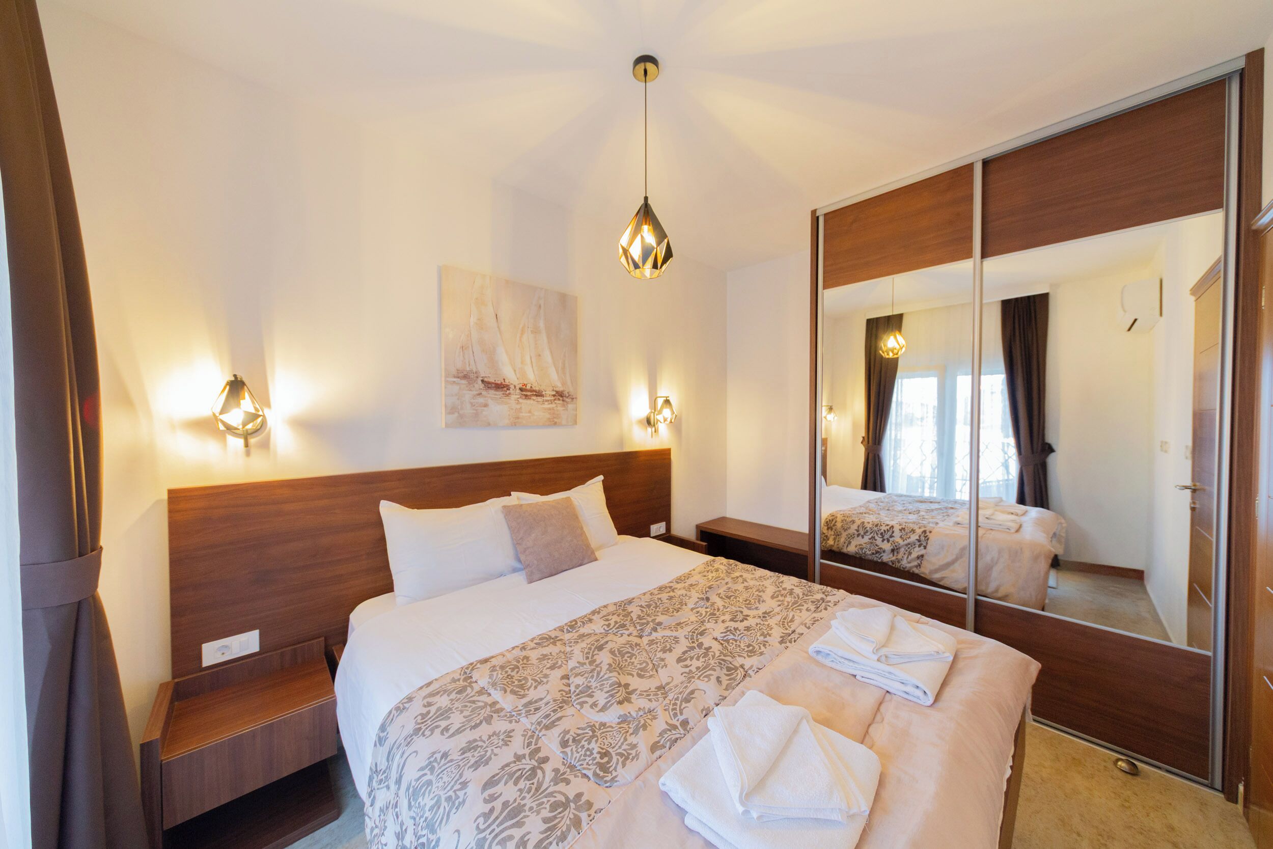 Une chambre d’hôtel avec un grand lit, une tête de lit en bois, une table de chevet, une lampe de chevet, un miroir et un luminaire fixé au mur.