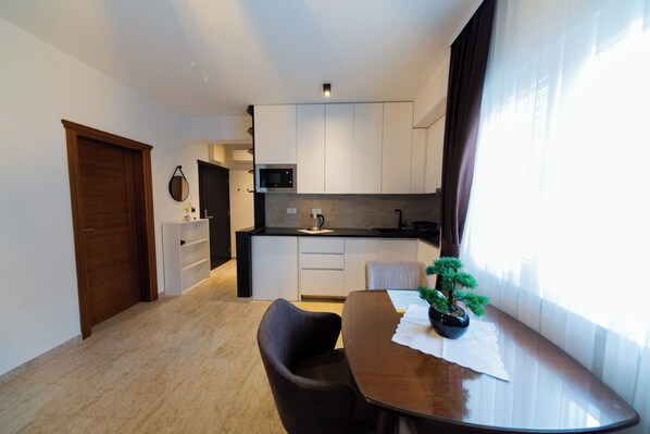 A modern kitchen with white cabinets, a microwave, and a sink. There is a dining table with a chair and a small potted plant.