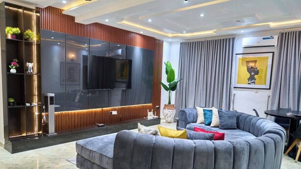 A modern living room with a large grey sectional sofa, a flat-screen TV, a built-in entertainment unit, and a dining area with a small table and chairs. - STBJ APARTMENT (Abuja)
