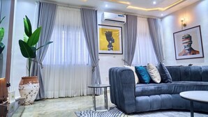 A modern living room with a grey sofa, a small round table, a potted plant, and a large window with grey curtains. - STBJ APARTMENT (Abuja)