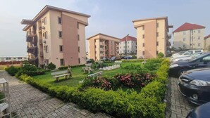 A residential building complex with multiple apartment buildings, a paved walkway, and a small garden area with benches and flowers. - STBJ APARTMENT (Abuja)