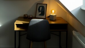 Een kleine, zwak verlichte kamer met een schuine plafond, een bureau met stoel, een klein tafeltje met lamp en een lijst met afbeelding aan de muur.