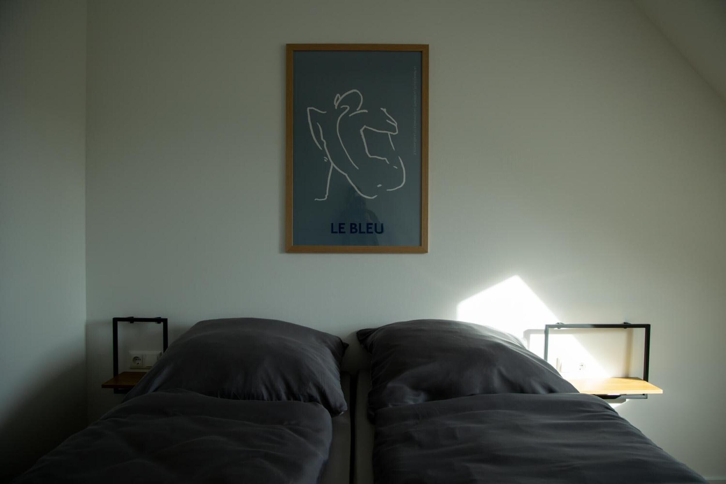 A bedroom with two beds, a framed artwork on the wall, and bedside tables with lamps.
