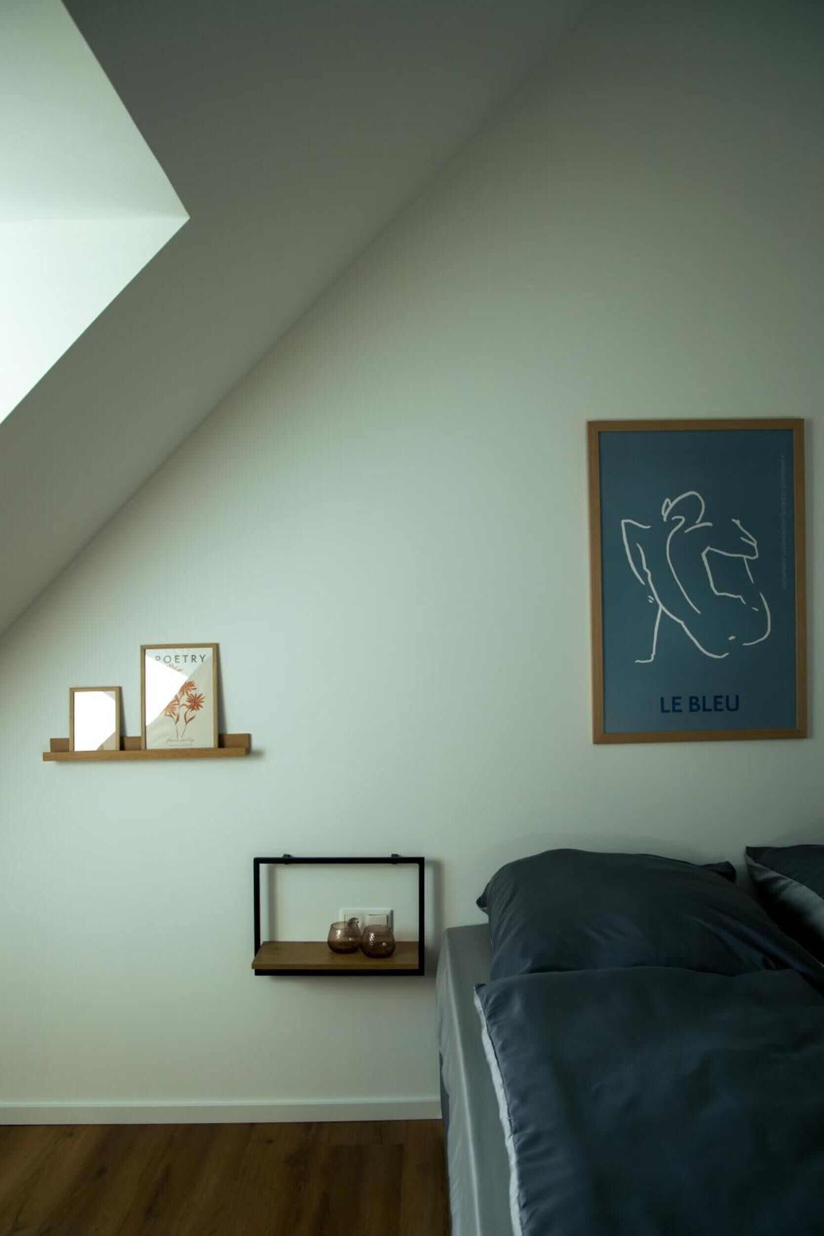 A bedroom with a sloped ceiling, a bed, a framed artwork, and two shelves with items.