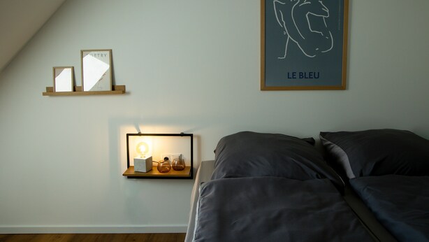 Een slaapkamer met een bed, een nachtkastje met een lamp en kunstwerken in een lijst aan de muur.