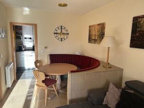 Een gezellig compacte ruimte met een eethoek, een bank, een lamp, een klok en een keuken op de achtergrond.