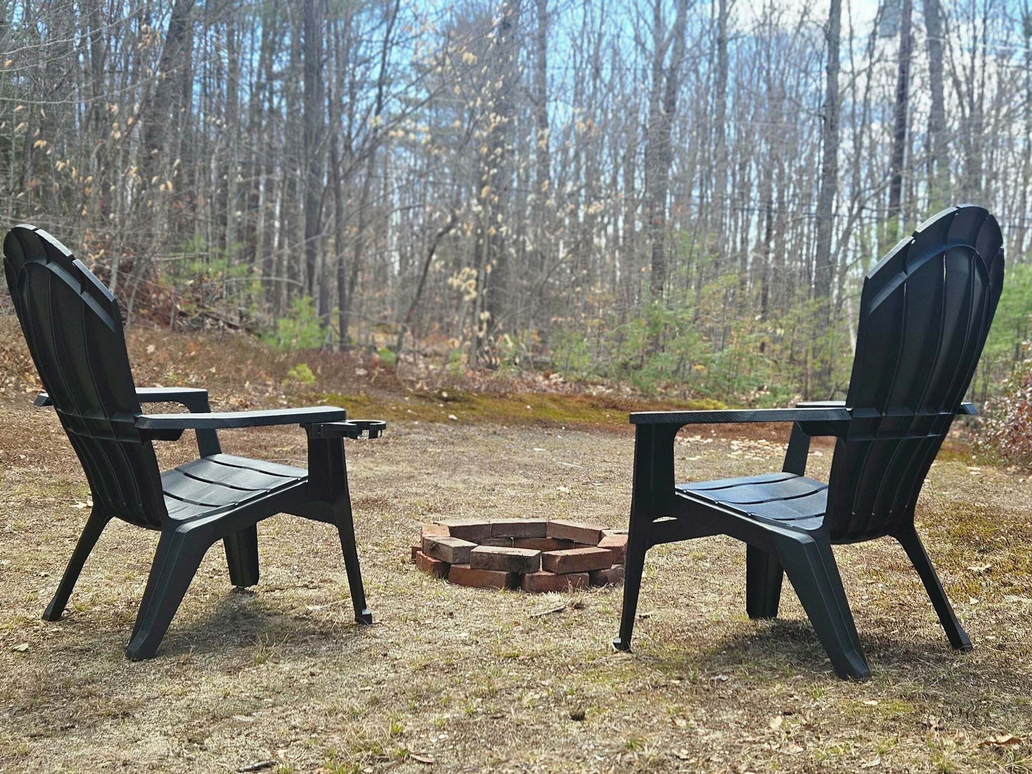 Two Adirondack chairs facing each other with a fire pit in between, set in a forested area.