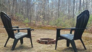 Two Adirondack chairs facing each other with a fire pit in between, set in a forested area.
