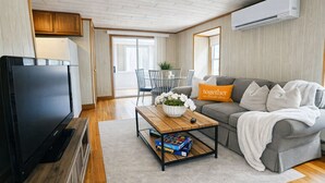 A modern living room with a flat-screen TV, a grey sofa with a 'together' pillow, a wooden coffee table with a plant and board games, and an air conditioning unit on the wall.