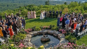 A wedding ceremony with guests seated on chairs arranged in a semi-circle around a stone fountain, set against a backdrop of a lush landscape with a lake and mountains.