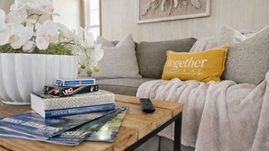 A cozy living room with a sofa, a coffee table with books and a remote control, a potted plant with white flowers, and a cushion with the text 'together'.