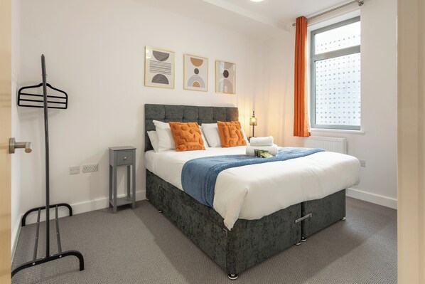A neatly made bed with a grey headboard, white bedding, and orange pillows. There are three framed artworks on the wall above the bed. - The Albany Sleeps 2 With Wifi (Redhill)