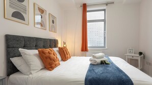 A neatly made bed with white and orange pillows, a blue blanket, and a small white nightstand with a clock and a plant. - The Albany Sleeps 2 With Wifi (Redhill)