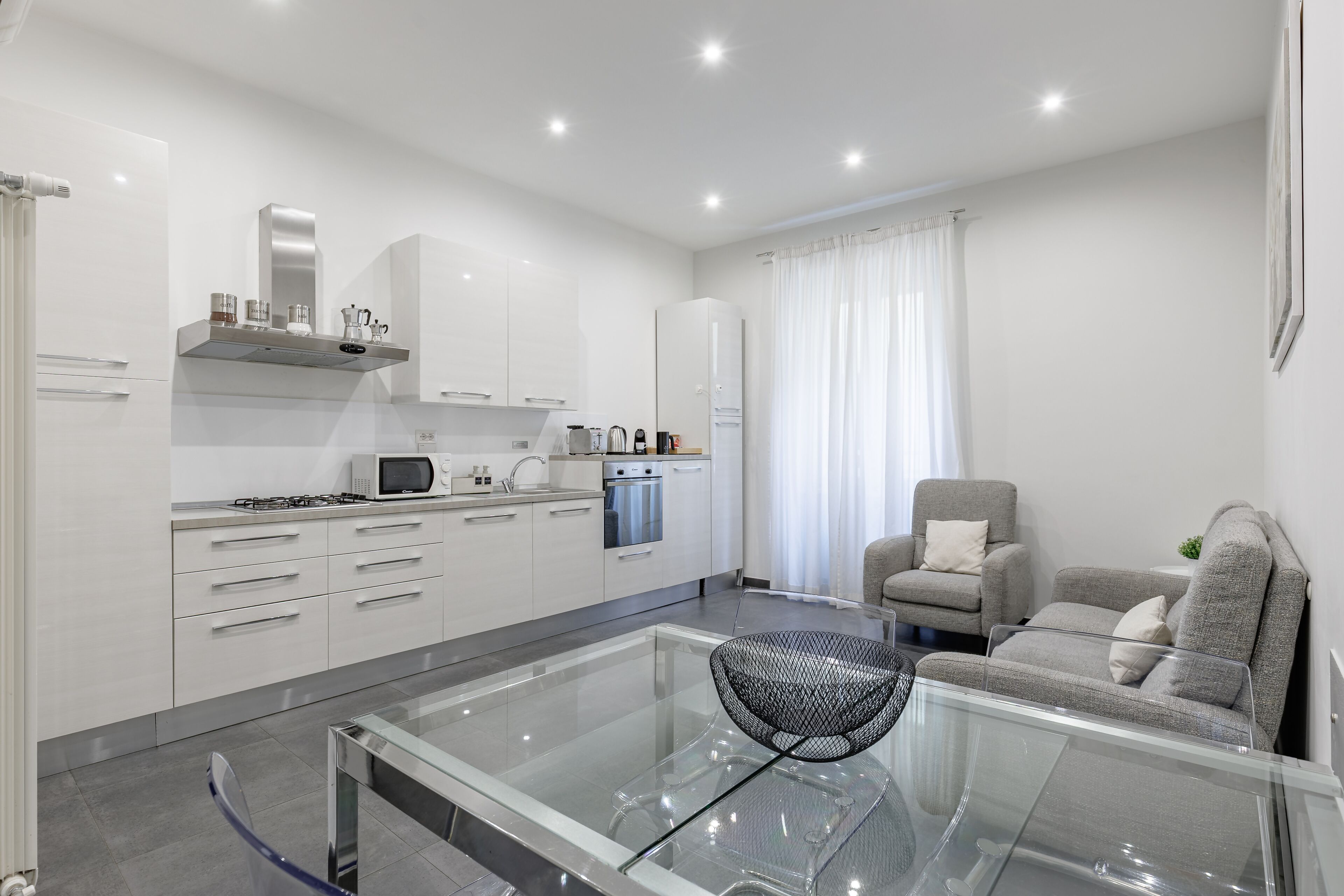 A modern kitchen with white cabinets, a glass dining table, and a grey sofa in a living area.