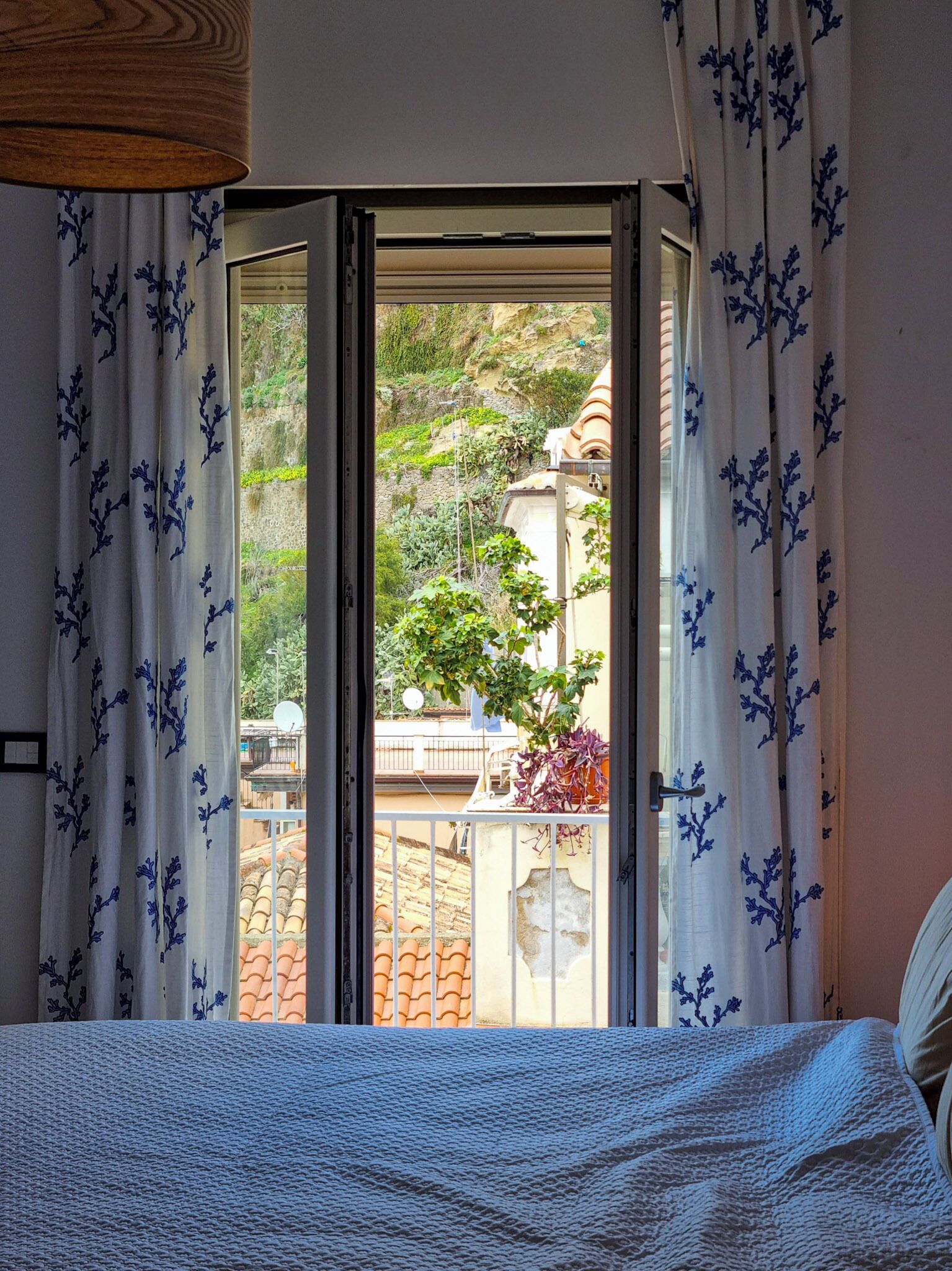 Ein Schlafzimmer mit Bett, einem Fenster mit Vorhängen und Blick auf einen Garten sowie Gebäude im Freien.