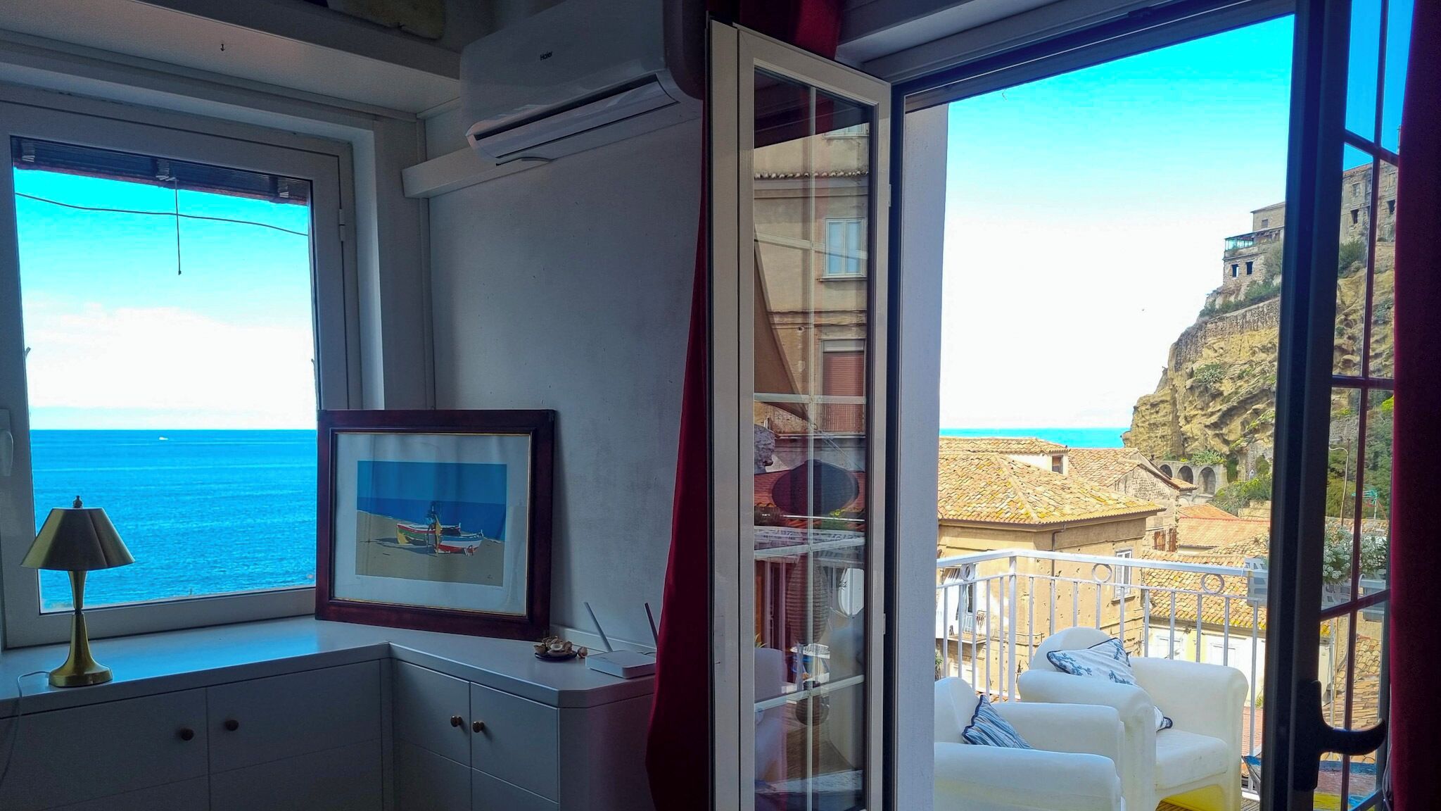 Ein Zimmer mit Meerblick, ein gerahmtes Bild auf einem Schreibtisch und ein Balkon mit weißen Stühlen.
