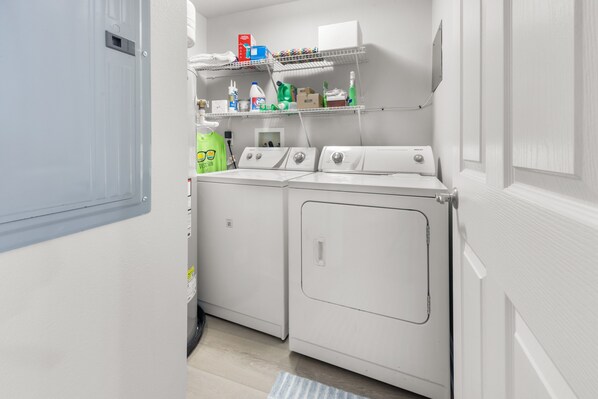 Uma lavanderia com máquina de lavar e secadora, prateleiras com produtos de limpeza e uma porta.