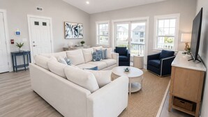 Una sala de estar con un sofá blanco, una mesa de centro redonda, un sillón azul y un televisor sobre un mueble.