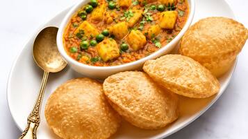 A bowl of curry with potatoes and peas, accompanied by three round, golden-brown bread items on a white plate.