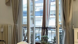 Een hotelkamer met een bed, gordijnen, een radiator en uitzicht op balkons en beplanting buiten.