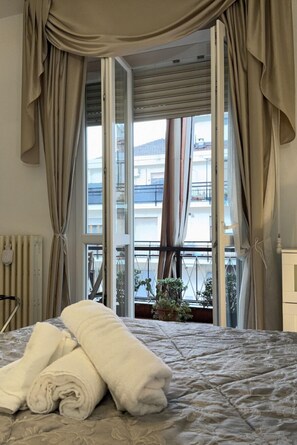 Ein Hotelzimmer mit Bett, Vorhängen, Heizkörper und Blick auf Balkone und Pflanzen.