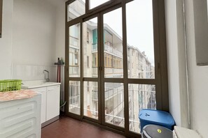 Eine schmale Küche mit einem großen Fenster, weißen Schränken, einem grünen Korb und einer blauen Mülltonne.