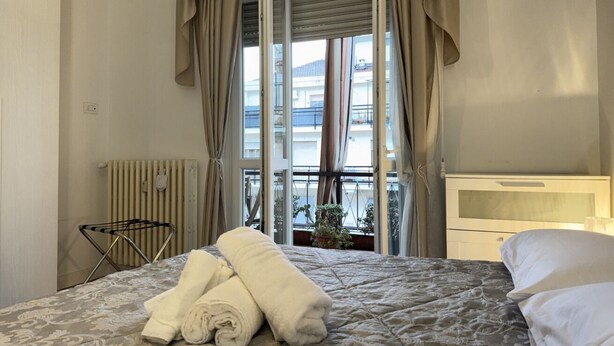 Hotelkamer met een bed, een radiator, een raam met gordijnen, uitzicht op gebouwen en een plant op de vensterbank.
