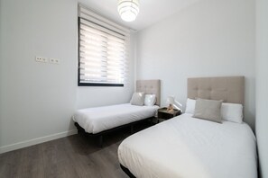 A hotel room with two beds, a window with blinds, a bedside table with a lamp, and a wall socket. - Comfortable apartment for families and business travelers   (Getafe)
