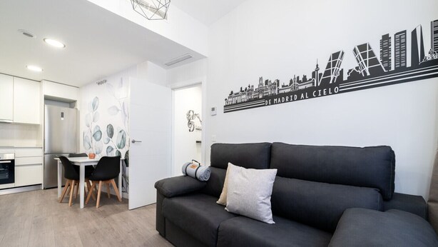 Una sala de estar moderna con un sofá negro, una mesita redonda y un comedor con una mesa y sillas blancas.