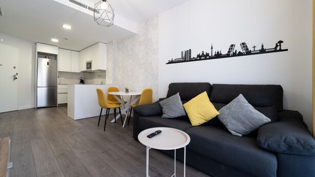Una sala de estar moderna con un sofá negro, una mesita redonda y un comedor con una mesa blanca y sillas amarillas.