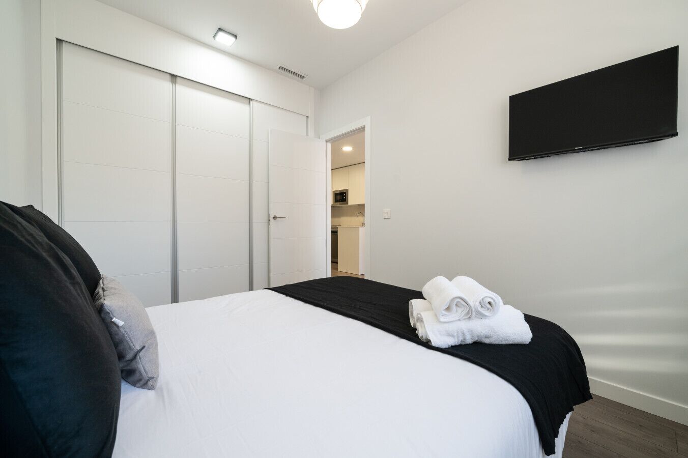 A hotel room with a bed, a flat-screen TV mounted on the wall, a door, and a view into the adjacent room.