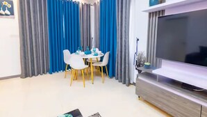 Ein modernes Wohnzimmer mit Essbereich, ausgestattet mit einem Flachbildfernseher, grauen Vorhängen und einem weißen Esstisch mit gelben Stühlen.
