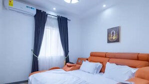 Hotellihuone, jossa on sänky, ilmastointilaite, ikkuna verhoilla ja seinään kiinnitetty valaisin.