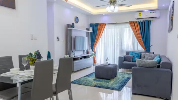 A modern living room with a grey sofa, a dining table with chairs, a TV, and a ceiling fan. - Colorful & Modern Escape: Pool, Balcony, Security! (Sakumono)