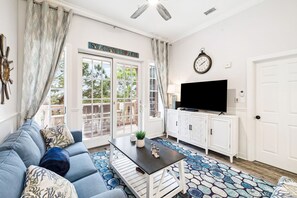 A living room with a sofa, coffee table, TV, and cabinets. There is a door with a sign above it and a clock on the wall. - Spacious Family Condo – Kid Friendly Stay w/ Bunk Room & Resort Style Amenities (Seacrest Beach)