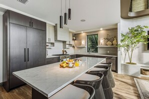 A modern kitchen with dark cabinets, a marble island, and a dining area with wicker chairs. - Calypso Current by Exclusive Vacations is a luxury new construction home in the (Anna Maria)