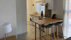 A modern kitchen with a central island, bar stools, a washing machine, and a large window with blinds. - Lovely flat, walk to Glentana beach... Play golf! (Great Brak River)