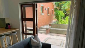 A room with a sofa, a dining table, and a view of a brick building and greenery outside. - Lovely flat, walk to Glentana beach... Play golf! (Great Brak River)