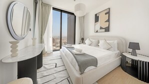A modern hotel room with a large bed, a round mirror, a bedside table with a lamp, and a view of the city through a large window. - Stylish 2BR + Maid’s Apartment in Sobha Hartland (Dubai)