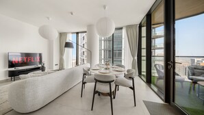 A modern living room with a large flat-screen TV, a round dining table, and a sofa. - Stylish 2BR + Maid’s Apartment in Sobha Hartland (Dubai)