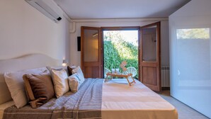 Ein Schlafzimmer mit einem Bett, Kissen, einem Nachttisch und Blick auf Grünanlagen durch eine offene Tür.