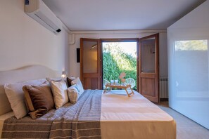 A bedroom with a bed, pillows, a bedside table, and a view of greenery through an open door. - ClickSardena apartment Terrace in Villa Fioretta with Pool and garden (Sardegna)
