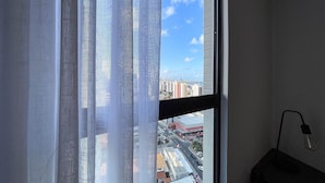 A window with sheer curtains, a view of a cityscape, and a lamp on the right. - Solaz by Hosppedar (João Pessoa)