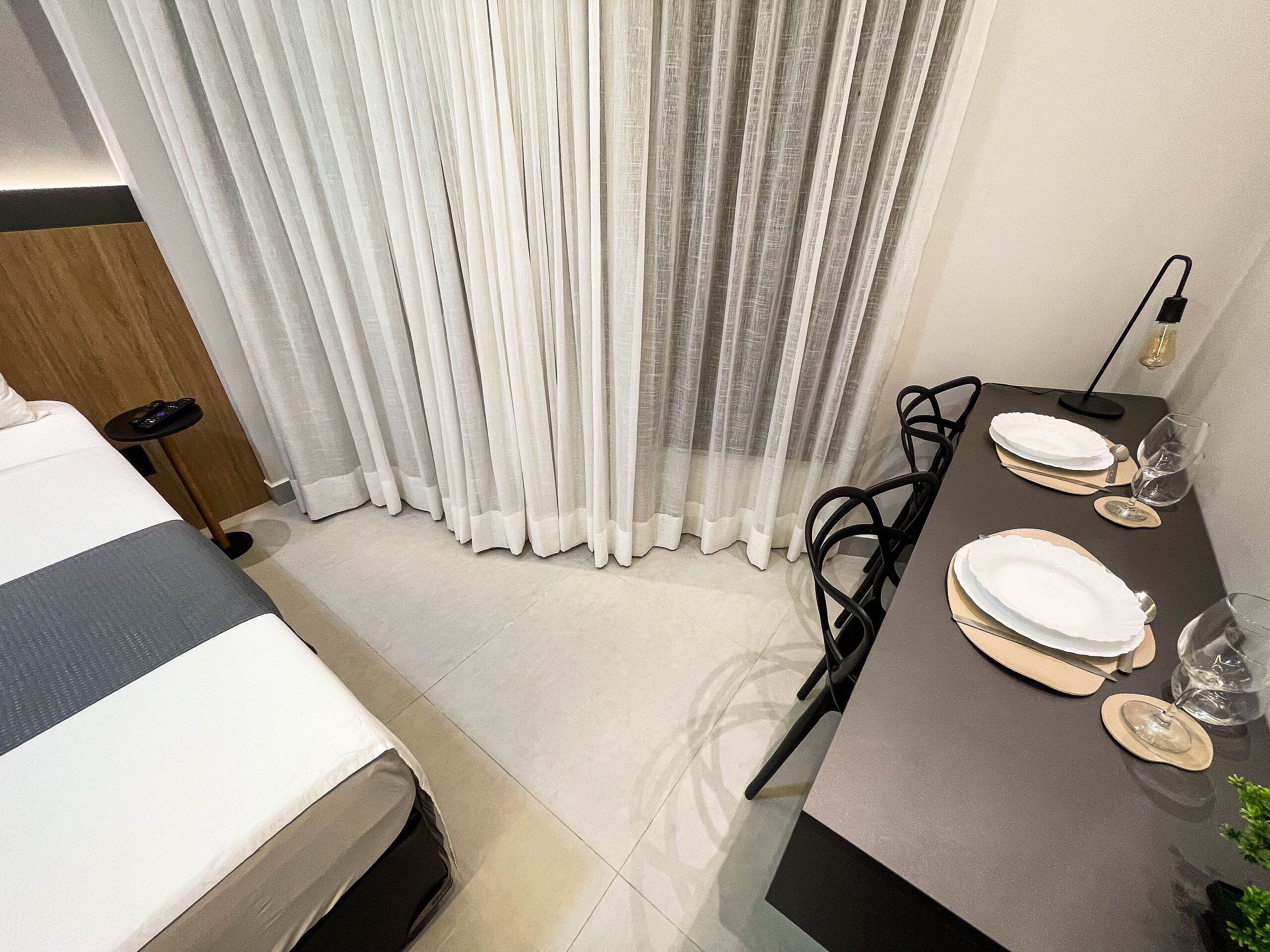 Habitación de hotel con cama, una mesa de comedor con platos y vasos, una silla y un control remoto sobre una mesita.