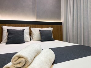 A neatly made bed with a white and dark gray color scheme, a wooden headboard, and a large window with curtains. - Solaz by Hosppedar (João Pessoa)