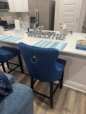 A modern kitchen with white cabinets, a stainless steel refrigerator, and a bar area with blue chairs and a 'Welcome' sign. - 3-bedroom townhome in superb Milton with cool AC (Milton)