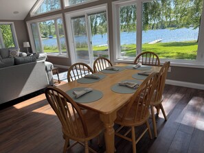 A dining area with a wooden table and chairs, large windows with a view of a lake, and a gray sofa in the background. - Perfect Waterfront Round Lake Haven with Clear Lake access, WiFi & AC (Fremont)